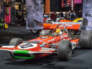 Een vintage rode raceauto met het nummer 10 en meerdere sponsorlogo's is binnen te zien bij Interclassics Maastricht, omringd door mensen en andere klassieke voertuigen op de achtergrond.