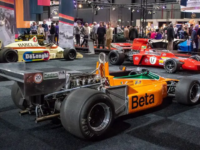 Verschillende vintage Formule 1-auto's worden tentoongesteld in een overdekte tentoonstellingshal bij Interclassics Maastricht, terwijl mensen op de achtergrond de voertuigen observeren.