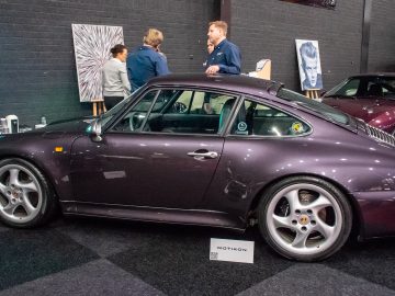 Bij Interclassics Maastricht staat een donkerpaarse Porsche 911 binnen, met drie mensen erbij. Op de achtergrond zijn schilderijen zichtbaar.