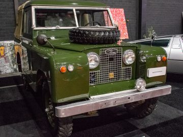 Binnen bij Interclassics Maastricht wordt een groene vintage Land Rover tentoongesteld, geflankeerd door andere auto's en kunstwerken aan de muur.