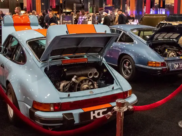 Twee vintage Porsche-auto's met de motoren achterin zichtbaar worden tentoongesteld op de indoor autoshow Interclassics Maastricht, omringd door een rode touwbarrière en bezoekers.