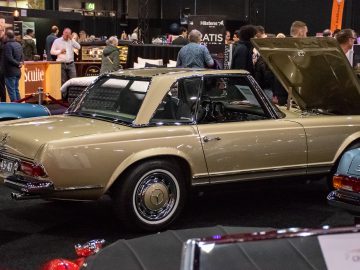 Op Interclassics Maastricht wordt een vintage beige Mercedes-Benz cabriolet met open kap tentoongesteld, omringd door aanwezigen.