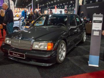 Op de Interclassics Maastricht staat binnen een zwarte Mercedes-Benz 190 E met een prijskaartje van € 198.000. Op de achtergrond zijn mensen te zien die geïnteresseerd zijn in de tentoonstelling.