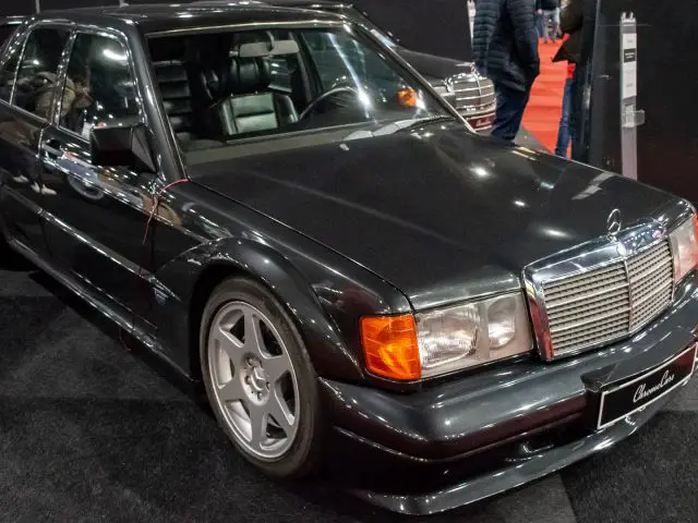 Een klassieke zwarte Mercedes-Benz 190E Evolution II wordt tentoongesteld op een vloerbedekking tijdens Interclassics Maastricht, omringd door andere voertuigen en mensen in een autotentoonstellingssetting.