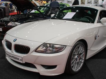 Op de gerenommeerde indoorautoshow Interclassics Maastricht schittert een witte BMW-sportwagen, terwijl op de achtergrond een andere zwarte auto met open motorkap zichtbaar is.