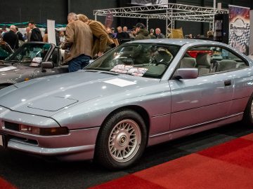 Een lichtblauwe BMW 8 Serie sportwagen wordt tijdens Interclassics Maastricht tentoongesteld op een rode loper, omringd door mensen en andere oldtimers.