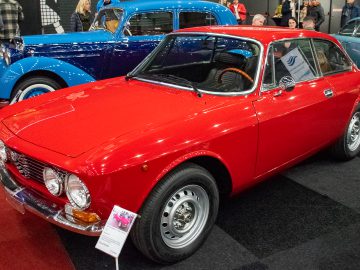 Op Interclassics Maastricht wordt een rode vintage coupé tentoongesteld naast andere klassieke auto's, met een bord voorop met gedetailleerde informatie over het voertuig.