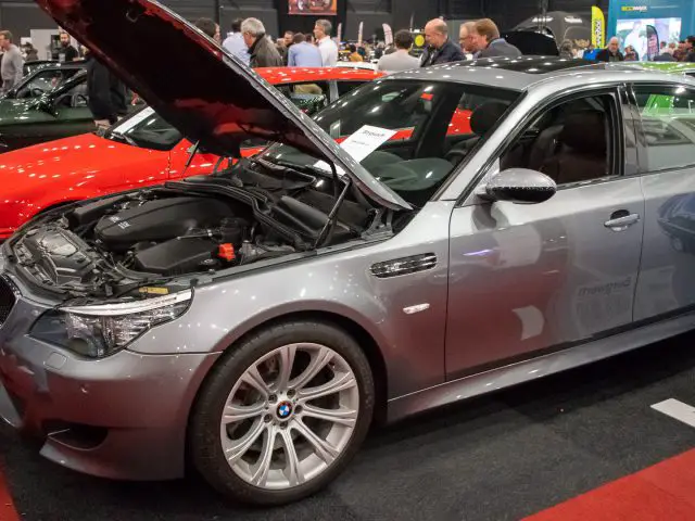 Een zilveren BMW met open kap wordt tentoongesteld op de indoor autoshow Interclassics Maastricht. Op de achtergrond zijn andere auto's en aanwezigen zichtbaar.