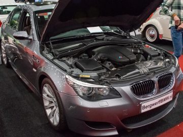 Op Interclassics Maastricht wordt een grijze BMW met open motorkap tentoongesteld, omringd door andere voertuigen in een indoor autoshow.