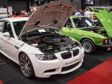 Bij Interclassics Maastricht staan twee auto's met open motorkap op een indoor autoshow. De witte auto is een BMW en de groene auto is een vintage BMW. Mensen staan er omheen en observeren de voertuigen.