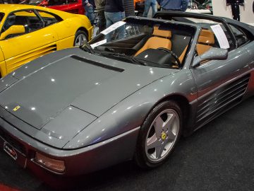 Bij Interclassics Maastricht staat een grijze Ferrari cabriolet met een lichtbruin interieur, omringd door bewonderaars en nog een gele Ferrari op de achtergrond.