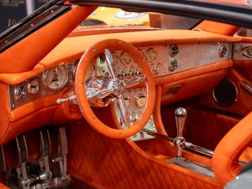 Het interieur van een auto met een oranje en chroomdesign, dat doet denken aan de klassieke schoonheden die werden tentoongesteld op Interclassics Maastricht. Hij beschikt over een stuurwiel, instrumentenpaneel en versnellingspook in vintage-stijl, terwijl de stoelen en het dashboard luxueus zijn bekleed met oranje leer.