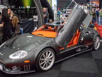 Op de autoshow InterClassics Maastricht wordt een strakke, grijze sportwagen met oranje interieur en geopende vlinderdeuren tentoongesteld, omringd door bezoekers en beursstands.