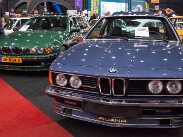 Een blauwe Alpina BMW en een groene BMW staan tentoongesteld op een indoor autoshow, met mensen op de achtergrond, tijdens Interclassics Maastricht.