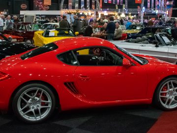 Op een drukke autoshow Interclassics Maastricht is een rode sportwagen te zien, omringd door andere klassieke en moderne auto's. Mensen lopen en onderzoeken de voertuigen.