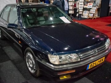 Een donkerblauwe Peugeot sedan met zichtbaar kenteken staat binnen geparkeerd op een rood-zwart geblokte vloer. Op de voorruit siert een te koop-bord, wat verwijst naar de verkrijgbaarheid ervan bij Interclassics Maastricht. Displayborden op de achtergrond dragen bij aan de sfeer van deze klassieke autotentoonstelling.