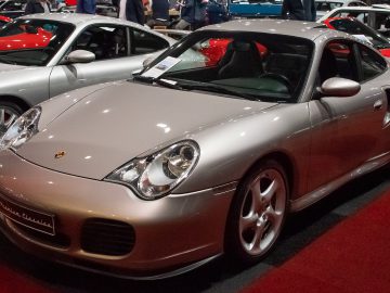 Een zilveren Porsche 911 wordt binnenshuis tentoongesteld op een rode loper bij Interclassics Maastricht, omringd door andere auto's, met een kaart op de voorruit.