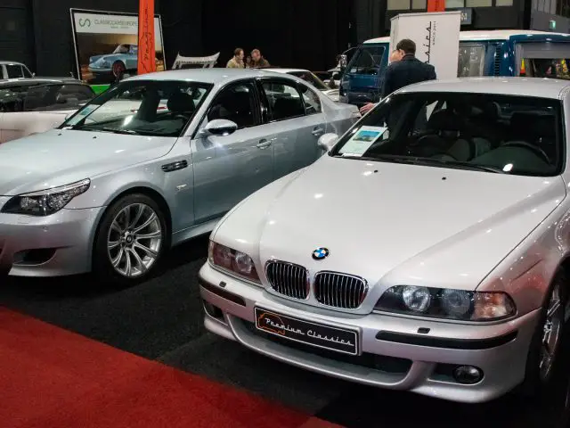 Twee BMW-auto's zijn binnen te zien op wat lijkt op de autoshow Interclassics Maastricht. Beide auto's zijn zilverkleurig en voor elk voertuig hangen informatieborden.