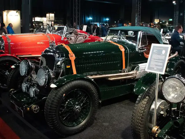 Een verzameling oldtimers, waaronder een donkergroene auto met leren banden, tentoongesteld op de indoor autoshow Interclassics Maastricht. Er staan mensen omheen, met informatieborden naast elke auto.