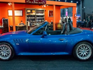 Binnen bij Interclassics Maastricht wordt een blauwe sportwagen met cabriolet tentoongesteld, met mensen en een bar op de achtergrond.