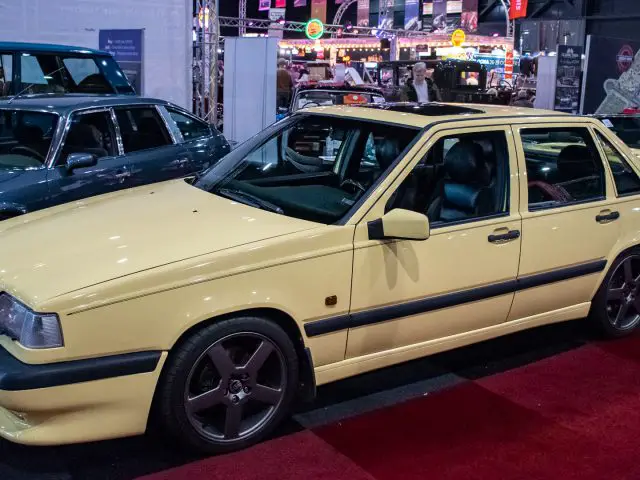 Op de indoor autoshow Interclassics Maastricht staat een gele klassieke sedan tentoongesteld, omringd door andere oldtimers en feestelijke verlichting.