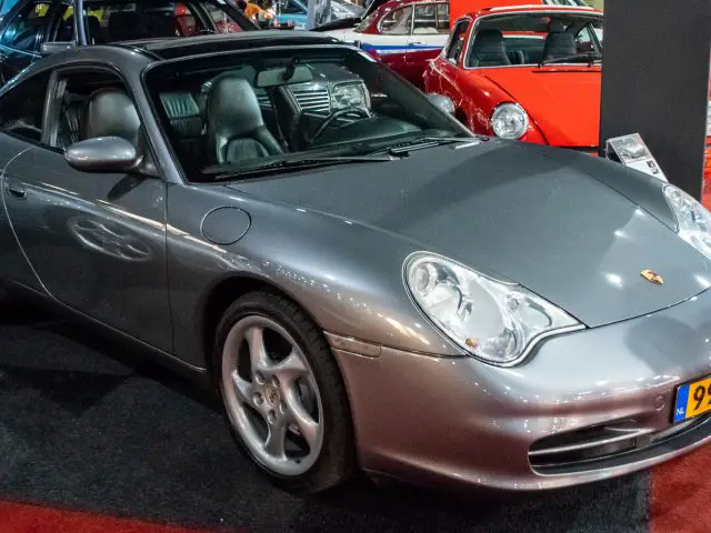 Een zilveren Porsche 911 Carrera staat elegant binnen op een rood-zwart geblokte vloer bij Interclassics Maastricht. Het kenteken is duidelijk zichtbaar en op de achtergrond zijn diverse andere auto's te zien.