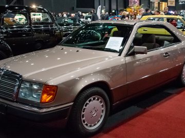 Op de autoshow Interclassics Maastricht wordt een beige Mercedes-Benz coupé tentoongesteld, met een informatiepapier op de voorruit. Op de achtergrond zijn andere klassieke auto's en mensen zichtbaar.