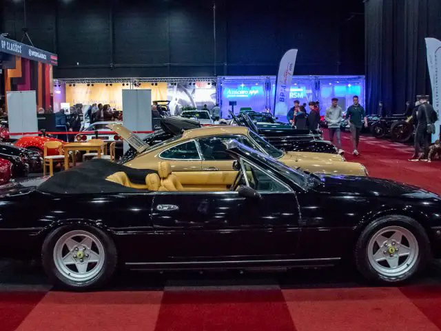 Een zwarte cabriolet met beige interieur wordt tentoongesteld op Interclassics Maastricht, een indoor autoshow met andere oldtimers, spandoeken en bezoekers op de achtergrond.