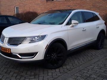 Een witte Lincoln MKX met getinte ramen en een Europees kenteken staat geparkeerd naast een bakstenen gebouw op een kasseistrook.
