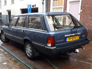 Een blauwe Peugeot 505 stationwagen geparkeerd op een natte geplaveide straat voor een bakstenen gebouw met verschillende borden en ramen. De auto heeft een geel Europees kenteken met de tekst "91-GTS-2.