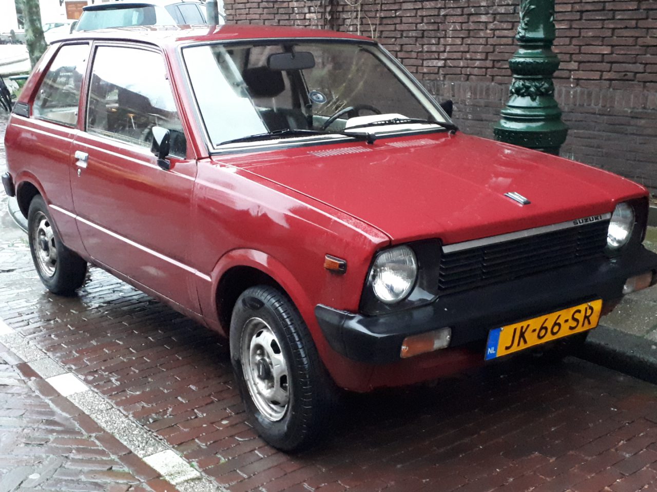 Een rode, compacte Suzuki Alto geparkeerd in een natte, geplaveide straat vlakbij een groene lantaarnpaal. De driedeurs auto heeft een geel kenteken met de tekst "JK-66-SR" en er zijn regendruppels zichtbaar op het oppervlak.