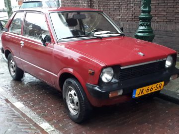 Een rode, compacte Suzuki Alto geparkeerd in een natte, geplaveide straat vlakbij een groene lantaarnpaal. De driedeurs auto heeft een geel kenteken met de tekst "JK-66-SR" en er zijn regendruppels zichtbaar op het oppervlak.