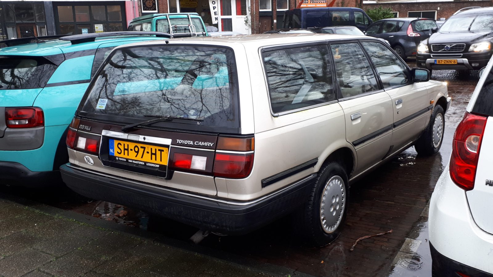 Een beige Toyota Camry stationwagen staat geparkeerd in een straat tussen een blauwe auto en een witte auto en valt op door zijn klassieke design.