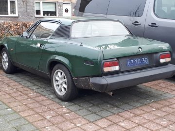 Een groene vintage Triumph TR7 coupé staat geparkeerd naast een grijs busje op een gedeeltelijk dichtgemetselde oprit. Op het kenteken van de auto staat ‘63-YA-30’.