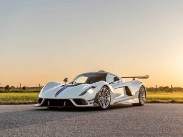 Een slanke, zilveren Hennessey Venom F5 Revolution Coupé met aerodynamische kenmerken staat geparkeerd op een weg tegen een prachtige zonsondergang op de achtergrond.