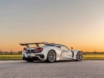 Een strakke witte Hennessey Venom F5 Revolution Coupé met een aerodynamisch ontwerp staat bij zonsondergang op een weg geparkeerd, voorzien van een grote achterspoiler en laagprofielbanden.