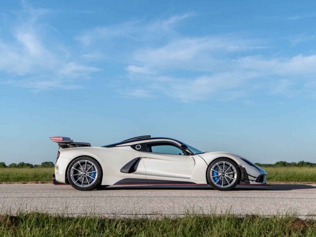 Een strakke, witte Hennessey Venom F5 Revolution Coupé met blauwe accenten staat geparkeerd op een open weg, tegen een achtergrond van helderblauwe lucht en groene velden.