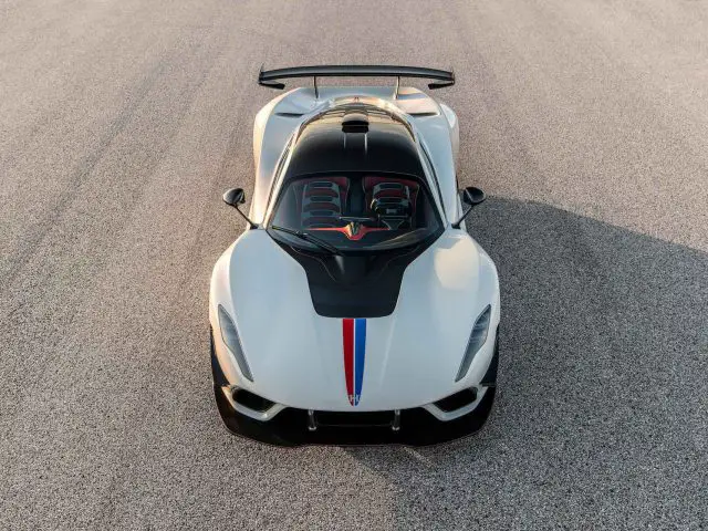 Een bovenaanzicht van een Hennessey Venom F5 Revolution Coupé, met zijn witte carrosserie, zwarte motorkap, prominente achtervleugel en rode en blauwe racestrepen, geparkeerd op een asfaltoppervlak.