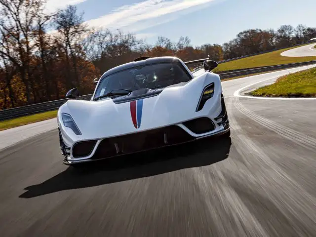 Een Hennessey Venom F5 Revolution Coupé, versierd met een rode en blauwe streep, rijdt op een heldere dag over een bochtige weg met bomen op de achtergrond.