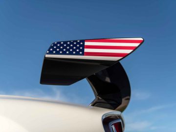 Een autospoiler van koolstofvezel met een embleem van de Amerikaanse vlag op de zijkant, tegen een helderblauwe hemel, vormt een aanvulling op het strakke ontwerp van de Hennessey Venom F5 Revolution Coupé.