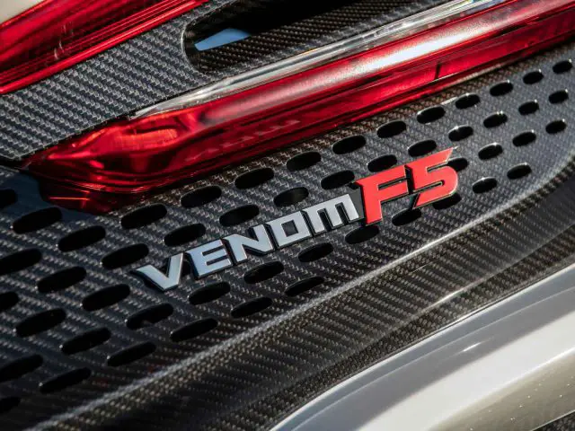 Close-up van de achterkant van een auto met het "Venom F5"-logo op een koolstofvezelachtergrond met een rood achterlicht erboven, wat het strakke ontwerp van de Hennessey Venom F5 Revolution Coupé benadrukt.