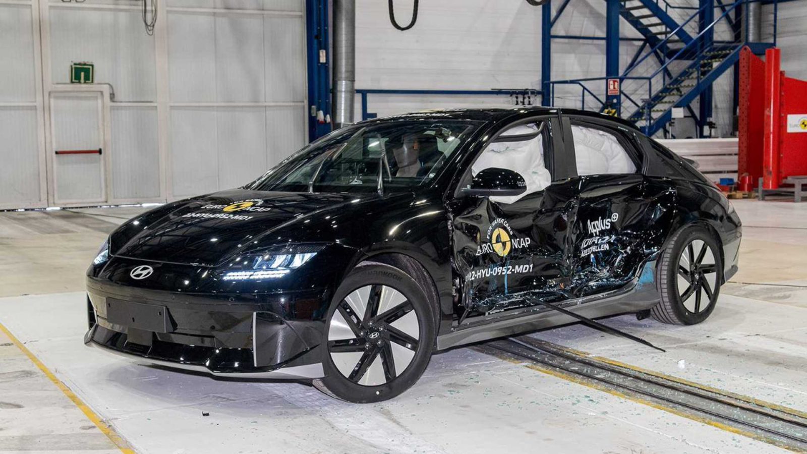 Een zwarte Ioniq 6 sedan met zichtbare zijschade ondergaat een crashtest in een industriële omgeving, met meerdere veiligheidstestlabels aan de buitenkant.