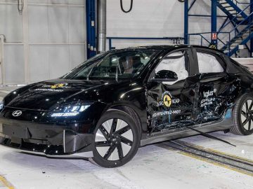 Een zwarte Ioniq 6 sedan met zichtbare zijschade ondergaat een crashtest in een industriële omgeving, met meerdere veiligheidstestlabels aan de buitenkant.