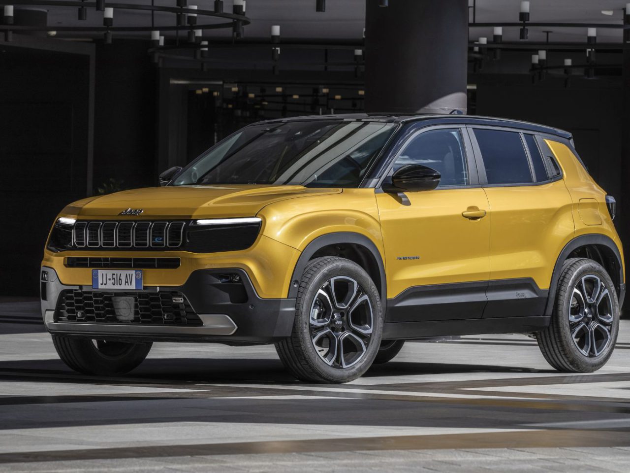 Een felgele Jeep Avenger SUV staat geparkeerd in een stedelijke omgeving met moderne architectuur op de achtergrond.