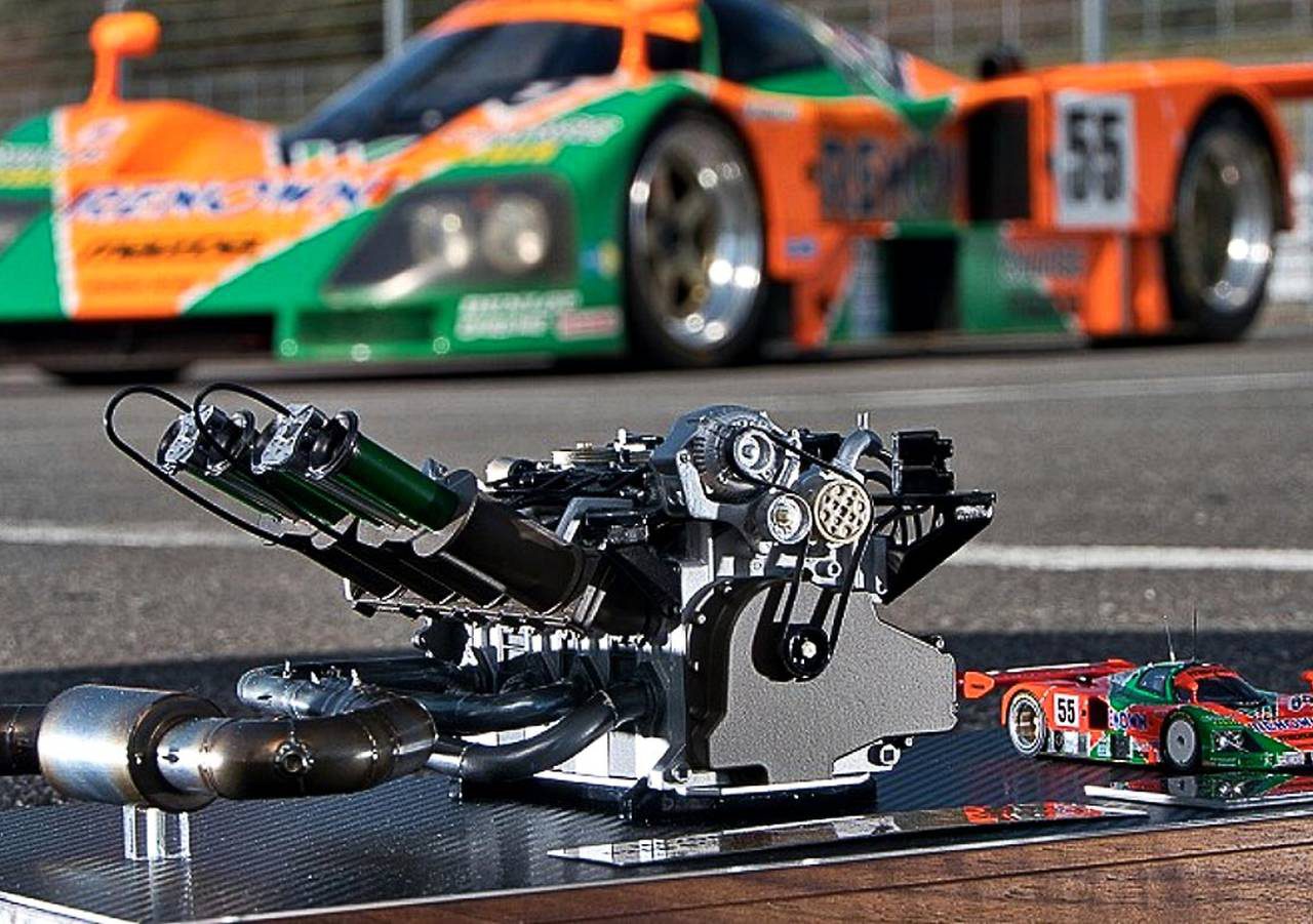 Close-up van een automotormodel weergegeven op een bord met een kleine gegoten auto, waarbij het ingewikkelde ontwerp van de wankelmotor wordt benadrukt. Een oranje en groene raceauto op ware grootte is wazig op de achtergrond.