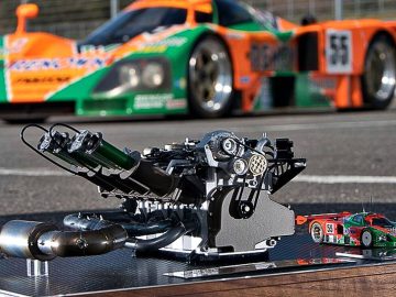 Close-up van een automotormodel weergegeven op een bord met een kleine gegoten auto, waarbij het ingewikkelde ontwerp van de wankelmotor wordt benadrukt. Een oranje en groene raceauto op ware grootte is wazig op de achtergrond.