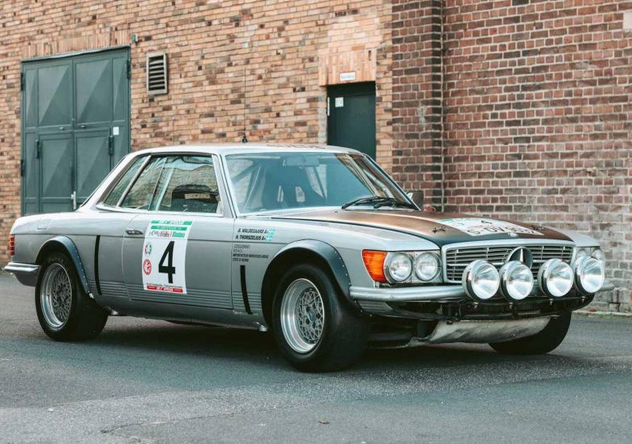 Een vintage zilveren Mercedes-rallyauto met het nummer 4 op de zijkant en vier schijnwerpers aan de voorkant, staat geparkeerd op straat tegen een bakstenen muur.