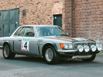 Een vintage zilveren Mercedes-rallyauto met het nummer 4 op de zijkant en vier schijnwerpers aan de voorkant, staat geparkeerd op straat tegen een bakstenen muur.