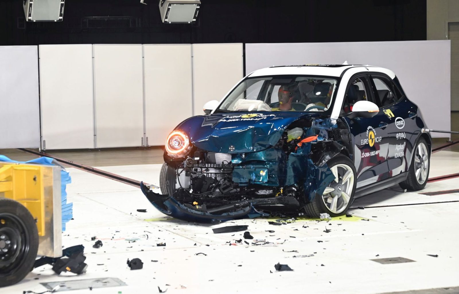 Een kleine Ora Funky Cat met aanzienlijke schade aan de voorkant ondergaat een crashtest in een gecontroleerde omgeving. Het puin ligt verspreid over het voertuig.