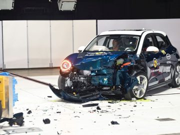 Een kleine Ora Funky Cat met aanzienlijke schade aan de voorkant ondergaat een crashtest in een gecontroleerde omgeving. Het puin ligt verspreid over het voertuig.